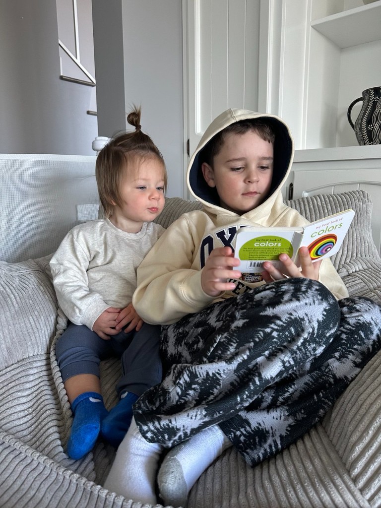 Aziz reading a picture book with a sibling on the couch